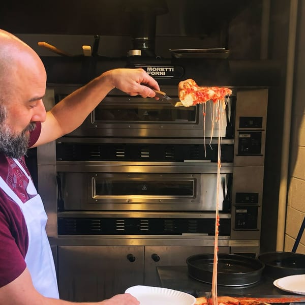 A chef pulls a slice of Campione's Italian pizza from a Moretti Forni oven, with cheese stretching.