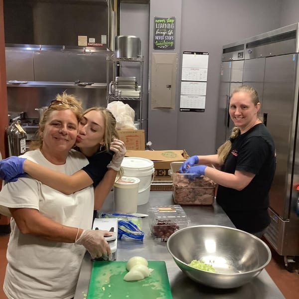 Three women in a commercial kitchen, two embracing, one preparing food, all wearing gloves.