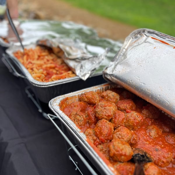 Italian meatballs in red sauce and pasta in foil catering trays on a black table.