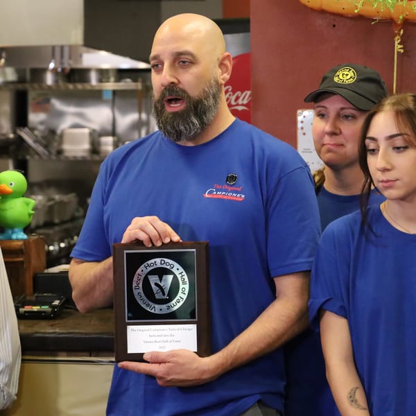 A man in a blue Campione's shirt holds a Vienna Beef Hall of Fame plaque, with two women beside him.